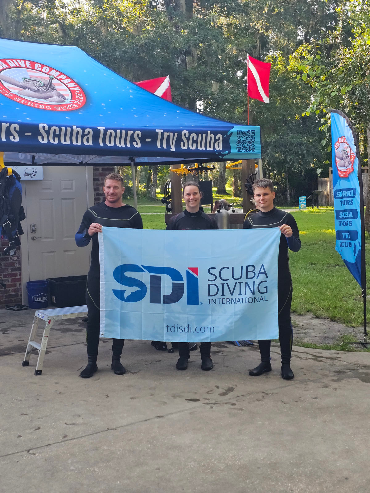 Three Try Scuba divers in wetsuits holding an SDI Scuba Diving International banner at the Florida Dive Company staging area